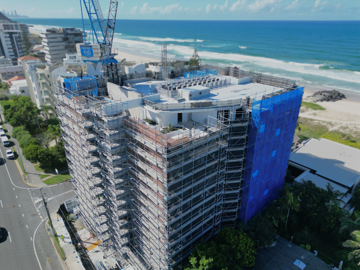 Construction of high-rise building near beach, blue netting, crane, scaffolding, sunny day.