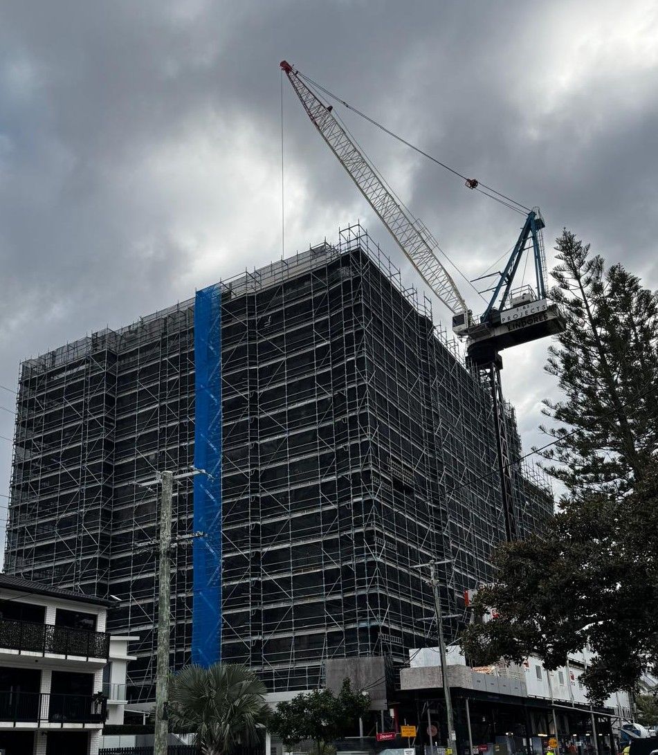 Construction of a tall high-rise building, vertical, partially completed, with a crane on top. Green and gray facade.