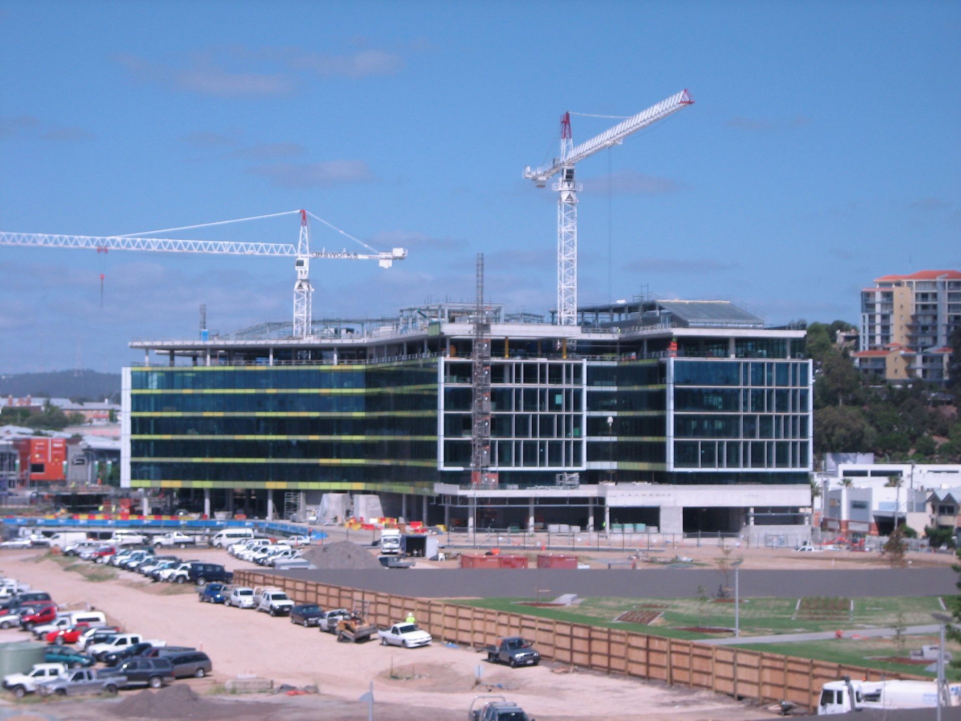 Construction site with cranes; modern building with glass and metal facade; cars parked in front.