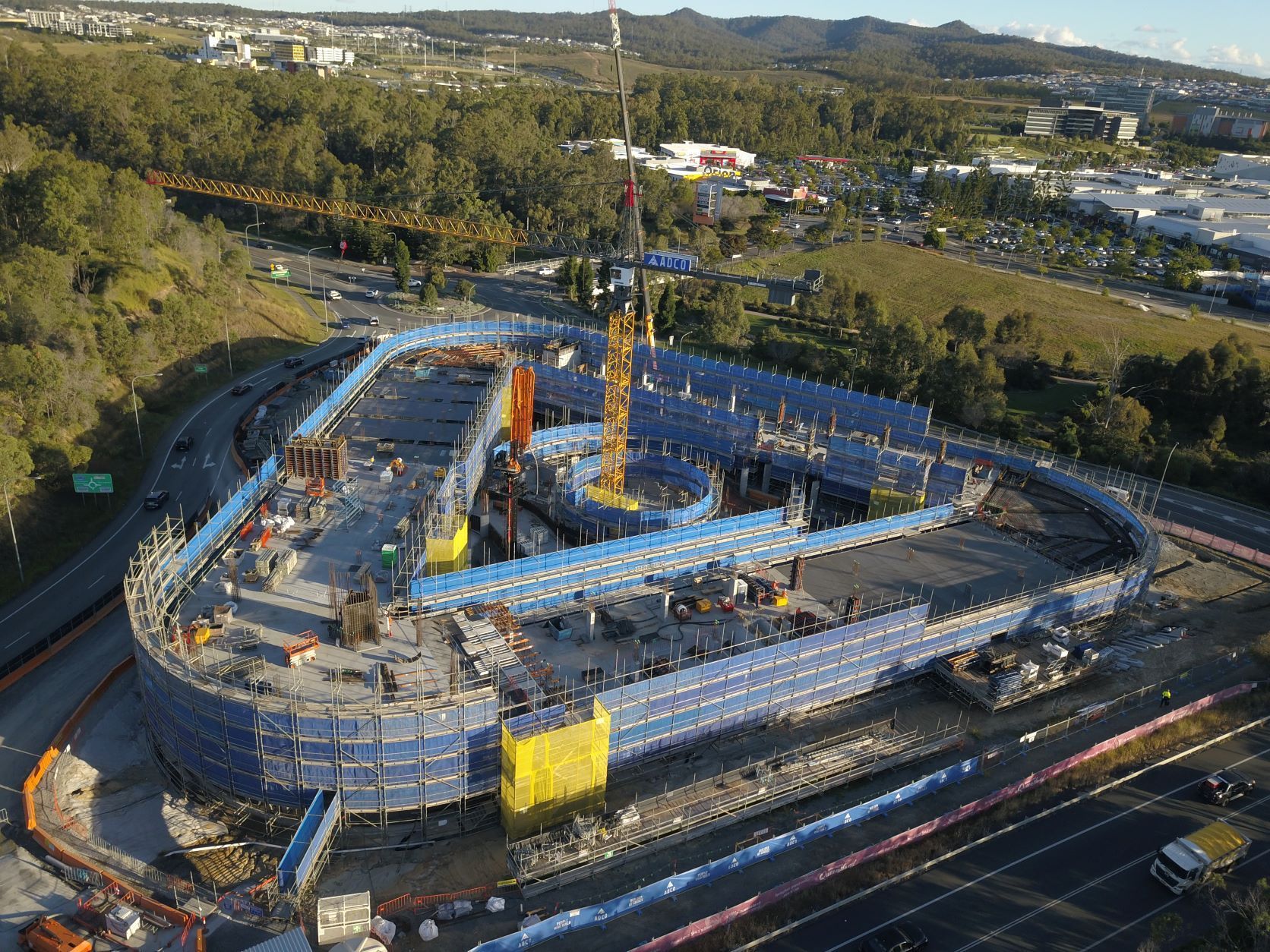 Construction site of a looping road overpass, with scaffolding and a crane surrounded by trees and a highway.