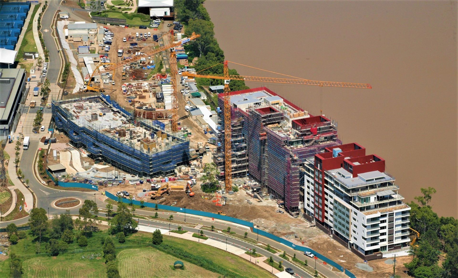 Construction site along a river; buildings in various stages, crane, vehicles, water.