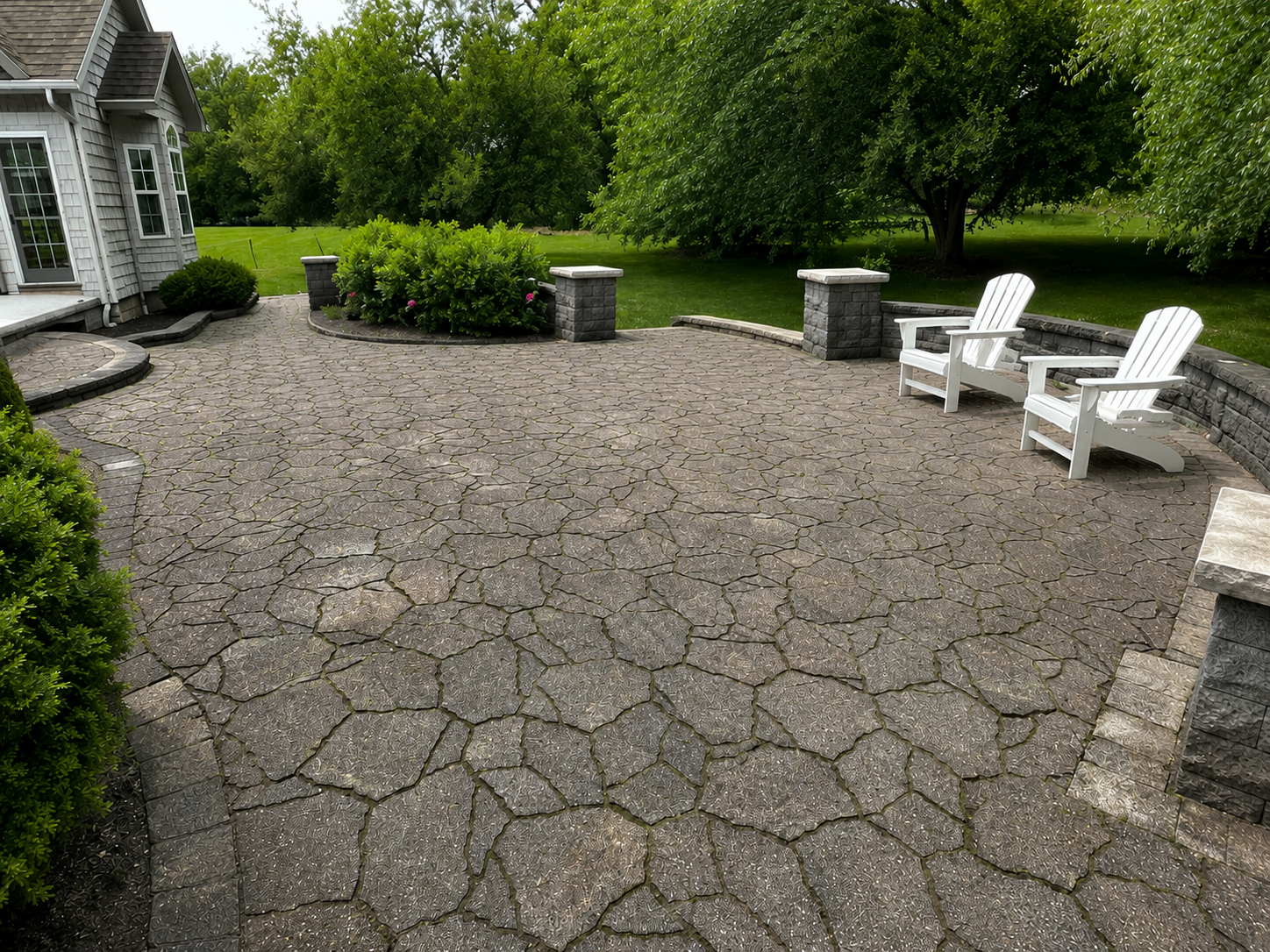 Stone patio with white chairs beside a house and green trees in a backyard