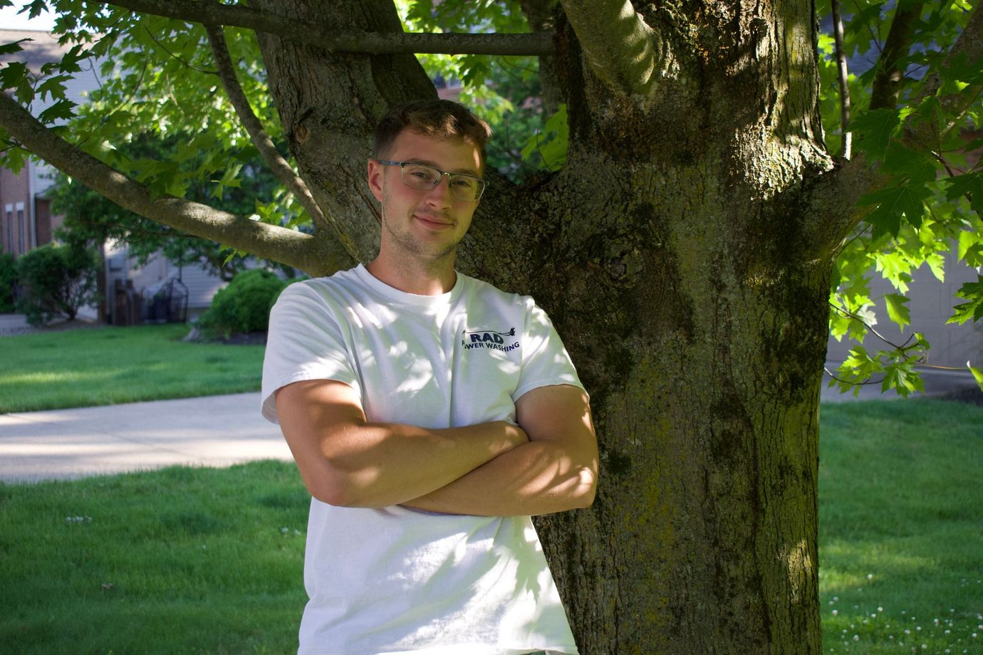 Person in a white T-shirt standing with arms crossed beside a tree in a sunny park