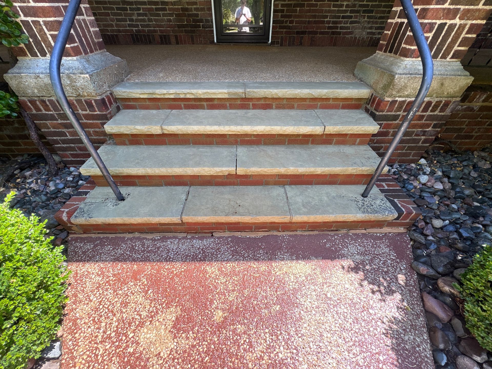 Front steps with blue railings leading to a brick porch, viewed over a red concrete walkway.