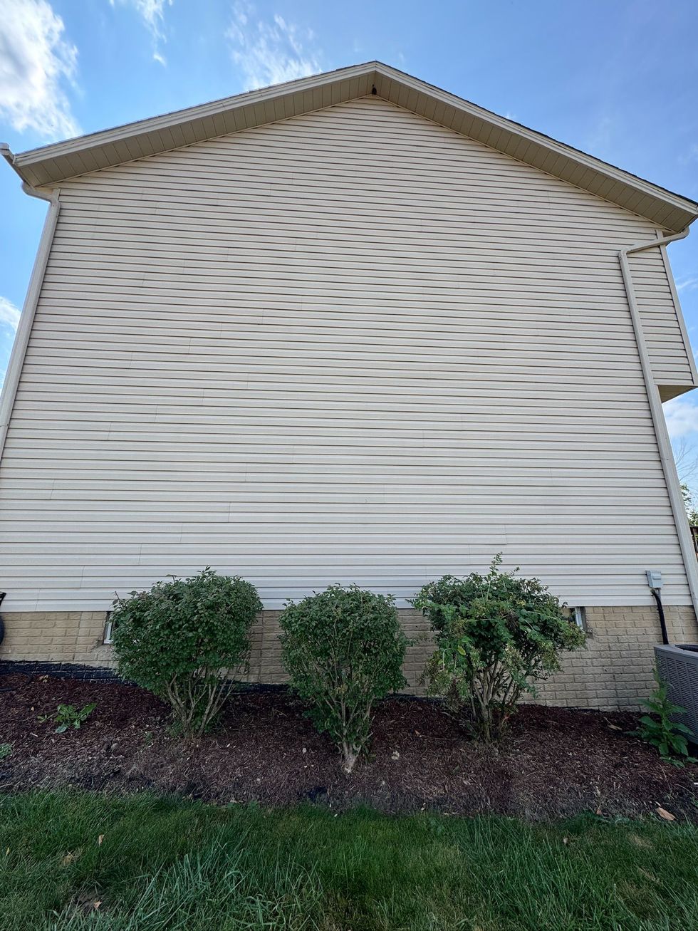 Side view of a beige house with vinyl siding, small shrubs, and a grassy lawn under a blue sky