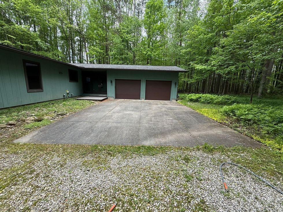 Gravel driveway leading to a two-car garage beside a green house, surrounded by trees.
