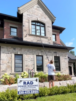 Worker pressure washing a stone house exterior beside a RAD Power Washing sign.