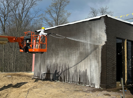 Worker pressure-washes the side of a dark brick building from an orange lift.