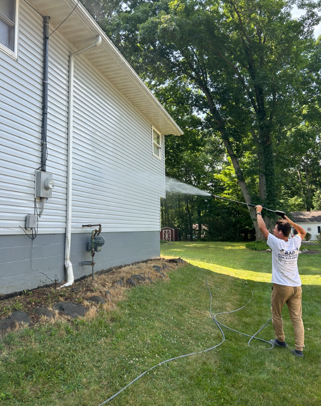 Person uses a long pole to work on the side of a white house in a grassy yard.