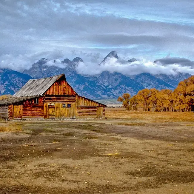Image of a rustic wooden barn in a wide field with snowy mountains and cloudy sky, ideal for gel finish treatment by Atlanta Canvas & Print