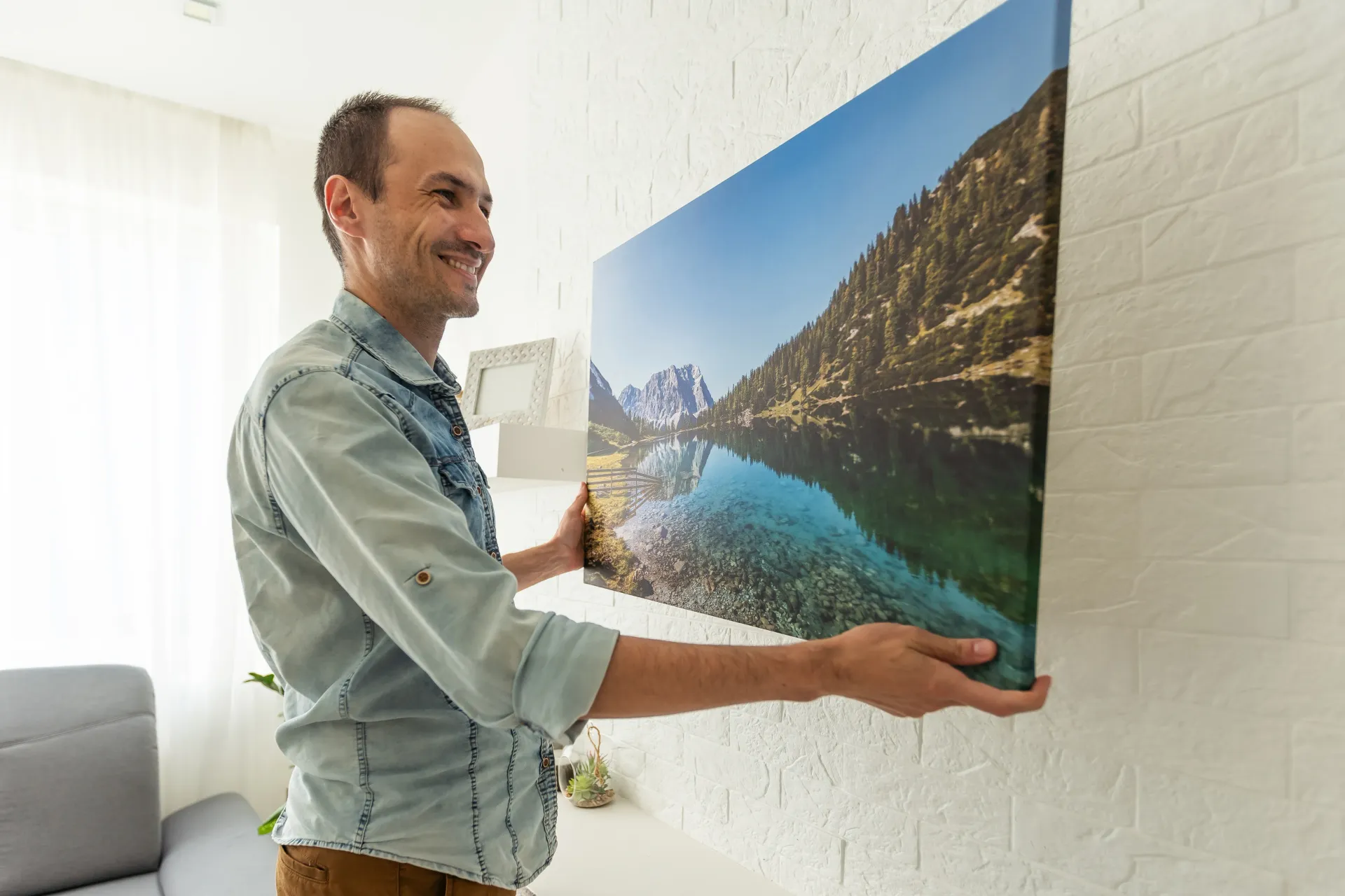 Man hanging a large landscape canvas print on a white wall in a bright room