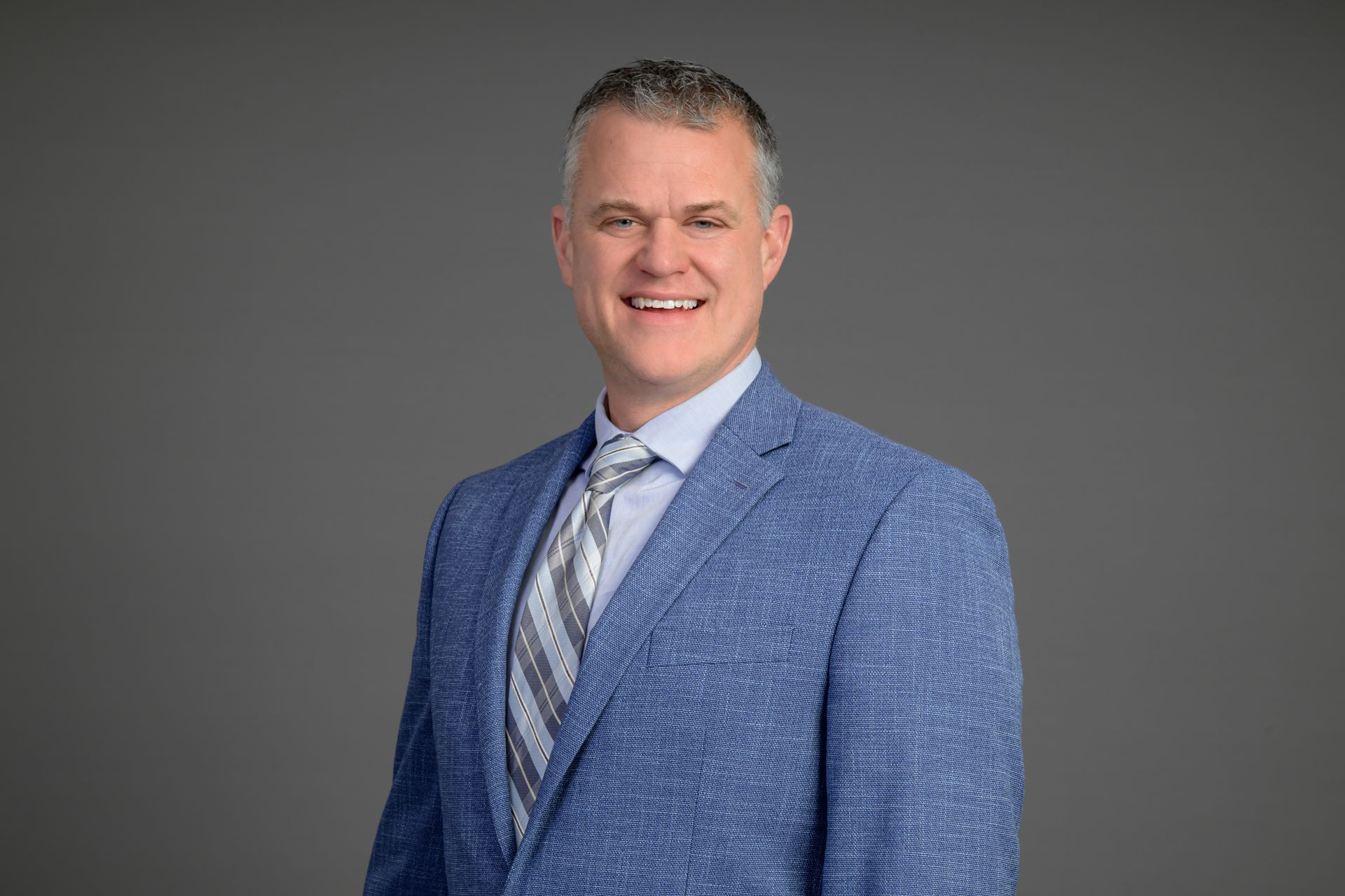 Man in a blue suit jacket and tie smiling against a gray studio background