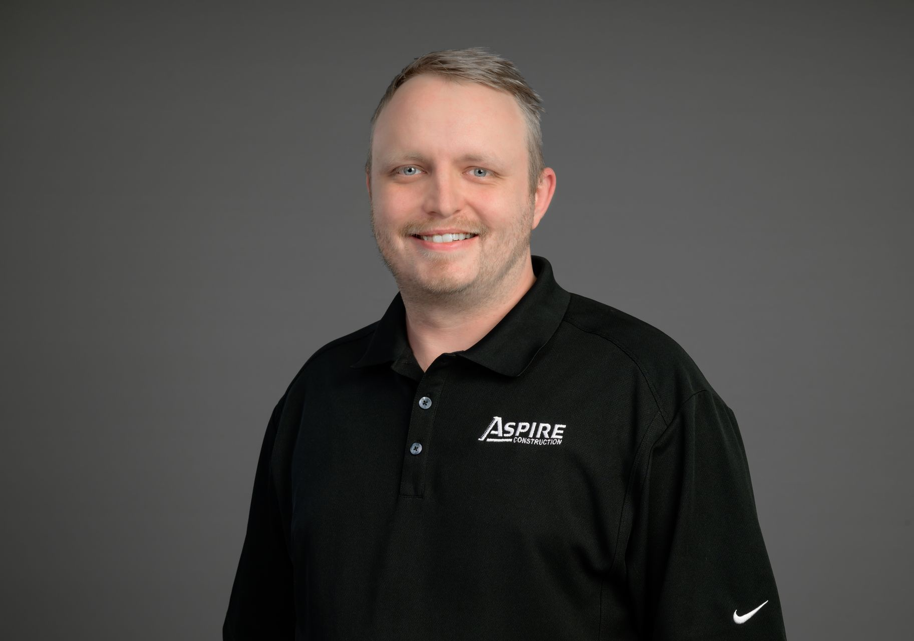 Smiling man in a black polo shirt with a logo, standing against a gray background