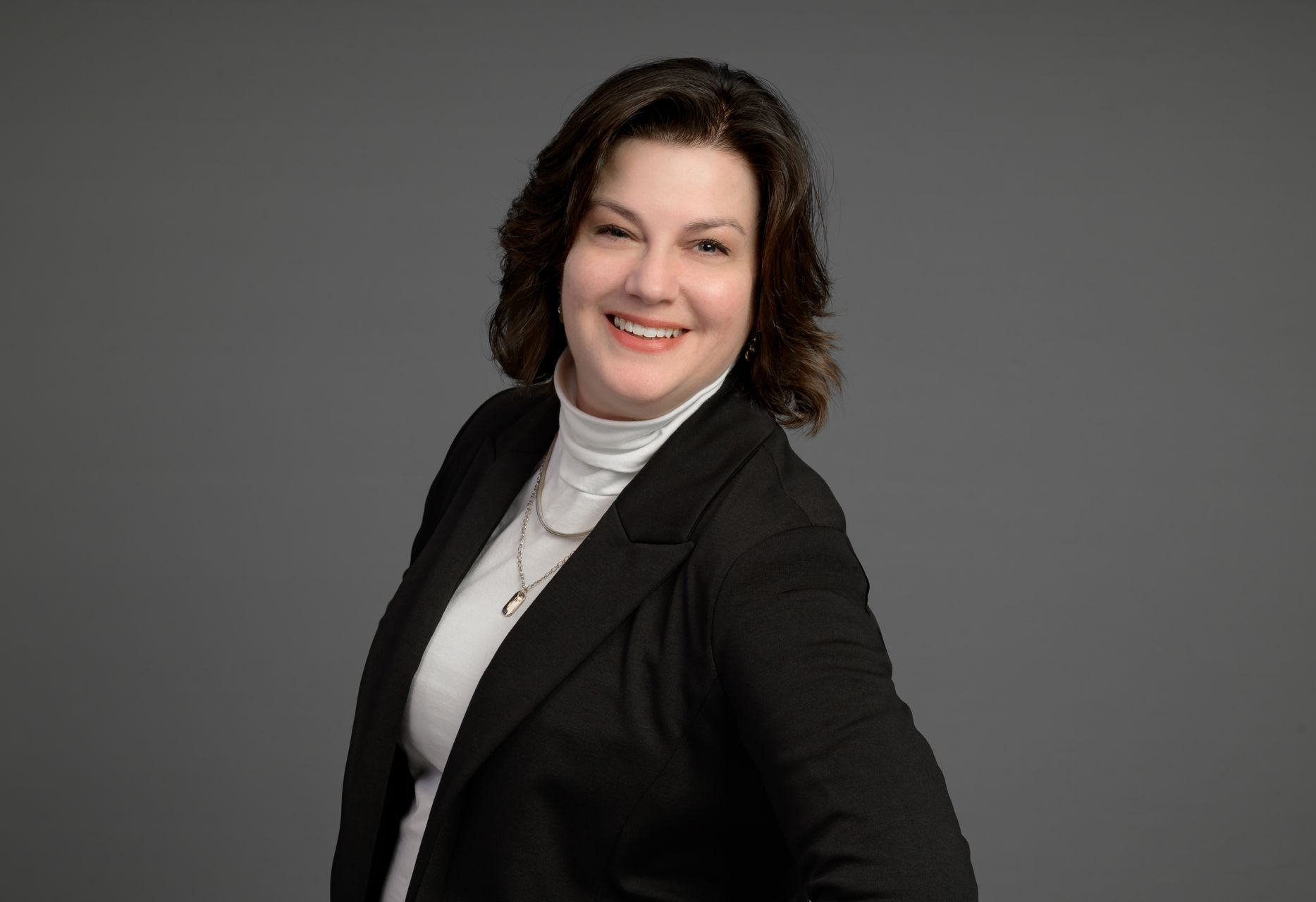 Smiling woman in a black blazer and white turtleneck against a gray background