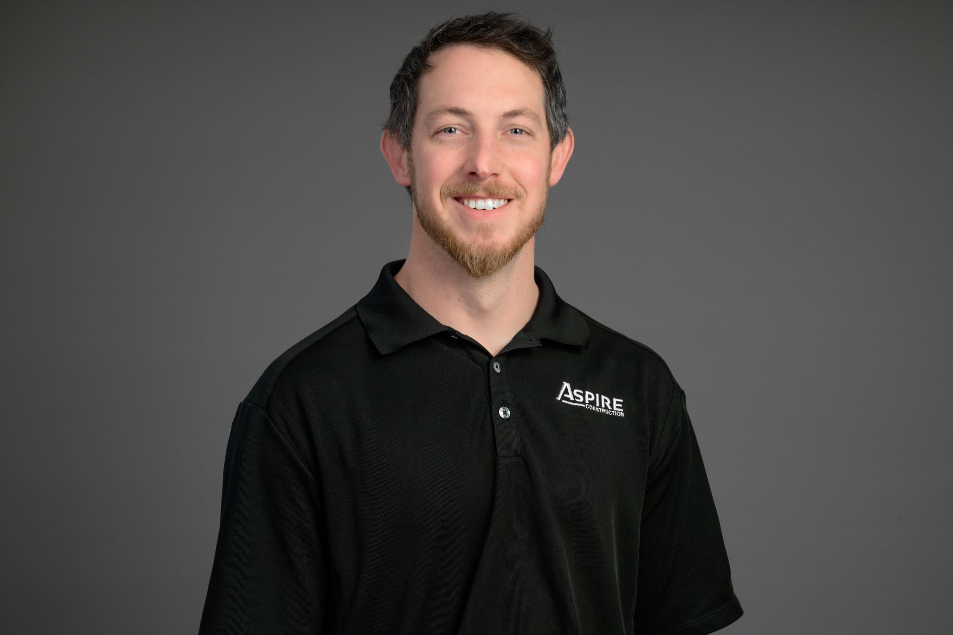 Smiling man in a black polo shirt with a logo, standing against a gray background