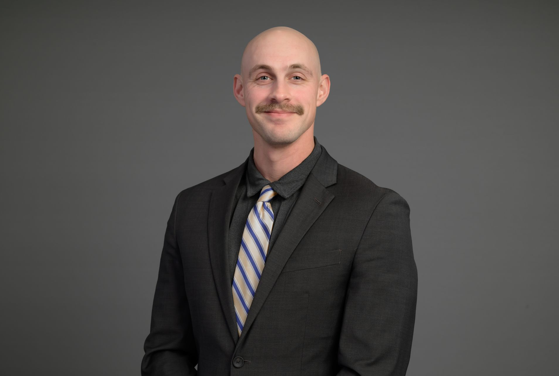 Portrait of a bald man with a mustache in a dark suit and striped tie against a gray background
