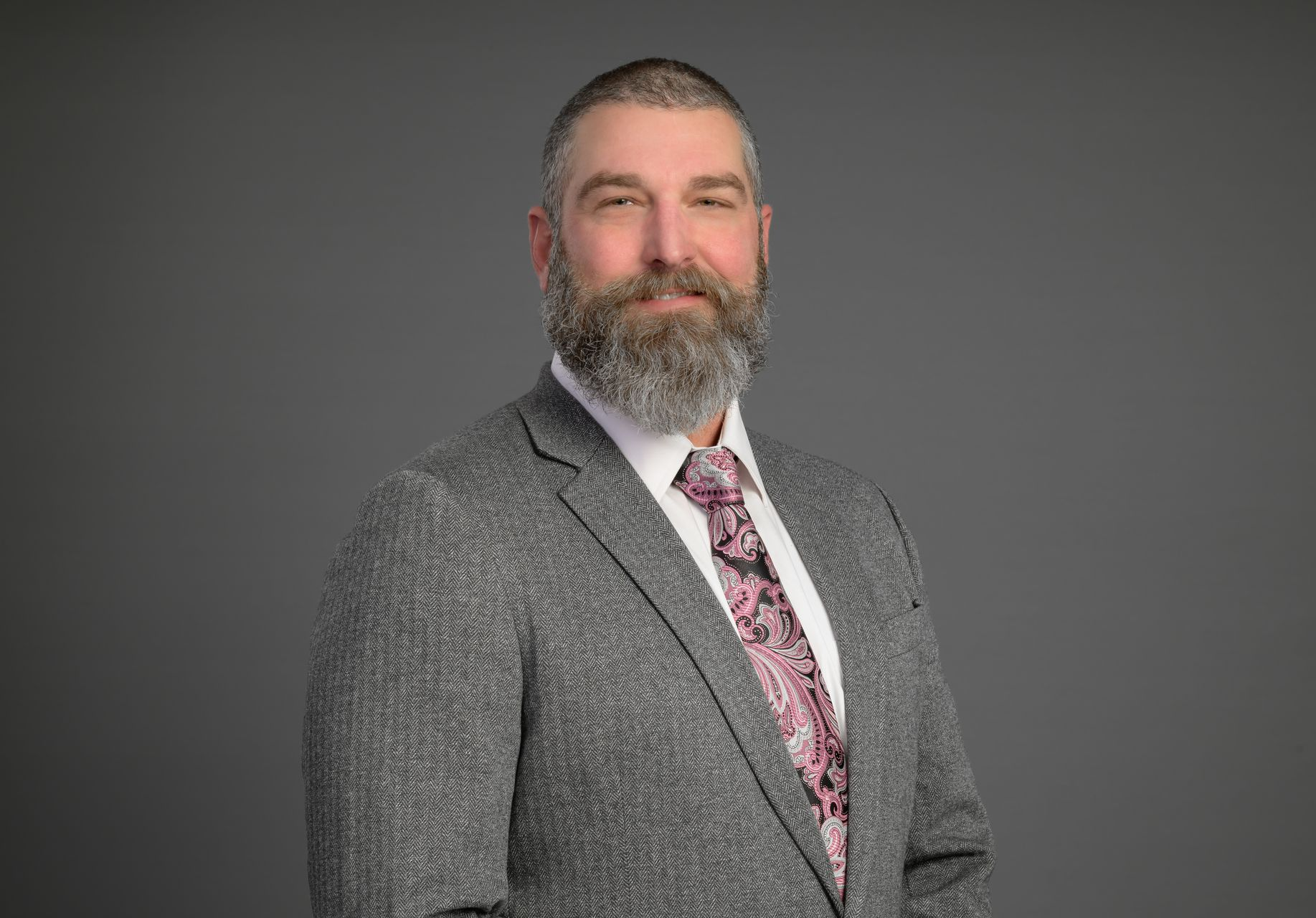 Studio portrait of a bearded man in a gray blazer and patterned tie against a gray backdrop