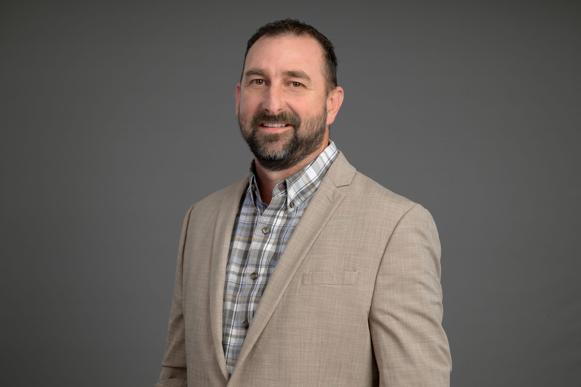 Portrait of a smiling man in a beige blazer and plaid shirt against a gray background