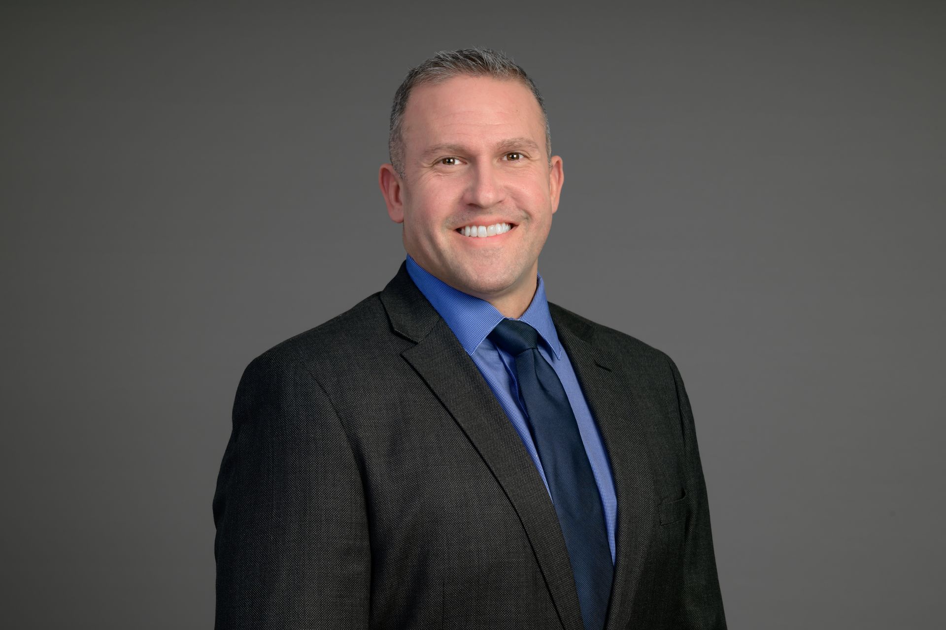Portrait of a smiling person in a dark suit, blue shirt, and striped tie against a gray background