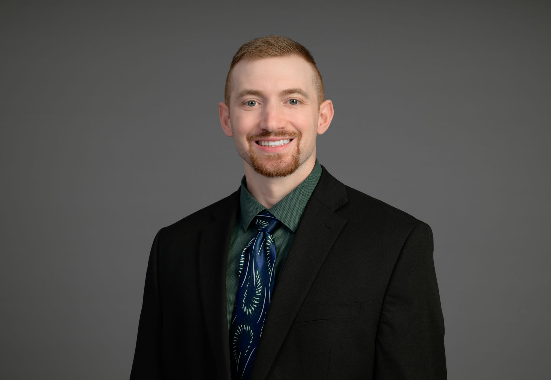 Smiling man in a black suit, dark shirt, and patterned tie against a gray background