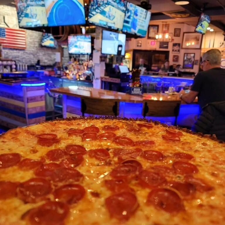 Pepperoni pizza on a table in a lively bar with blue-lit counters and TV screens￼