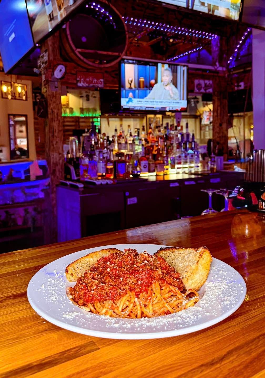 Plate of pasta with garlic bread on a wooden bar in a lively sports bar with TVs and purple lighting