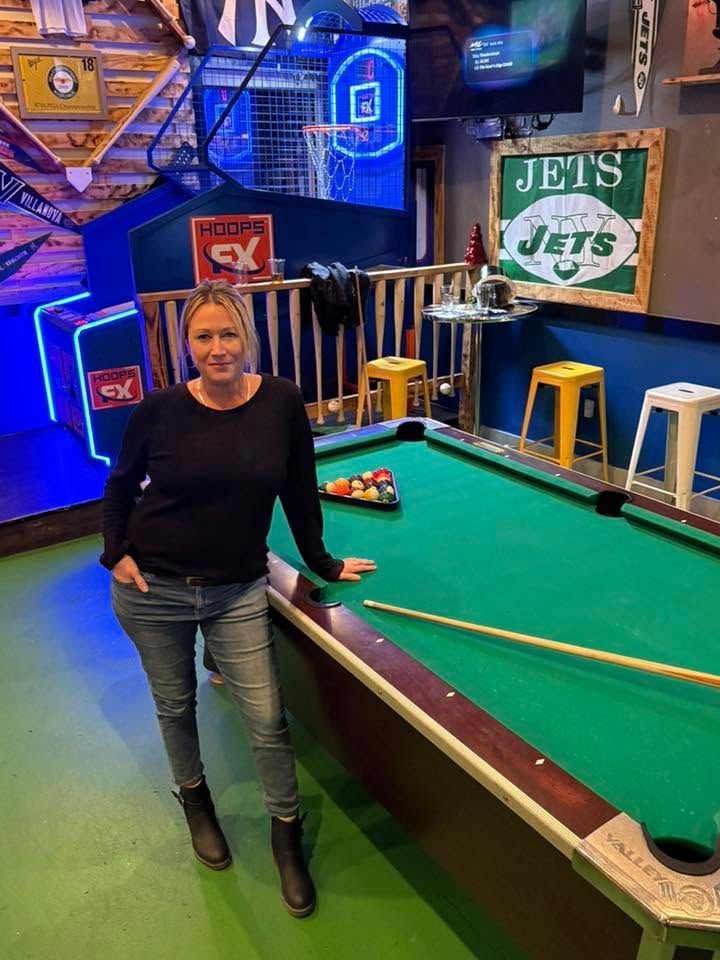 Woman leaning on a pool table in a sports bar with neon signs and New York Jets decor.