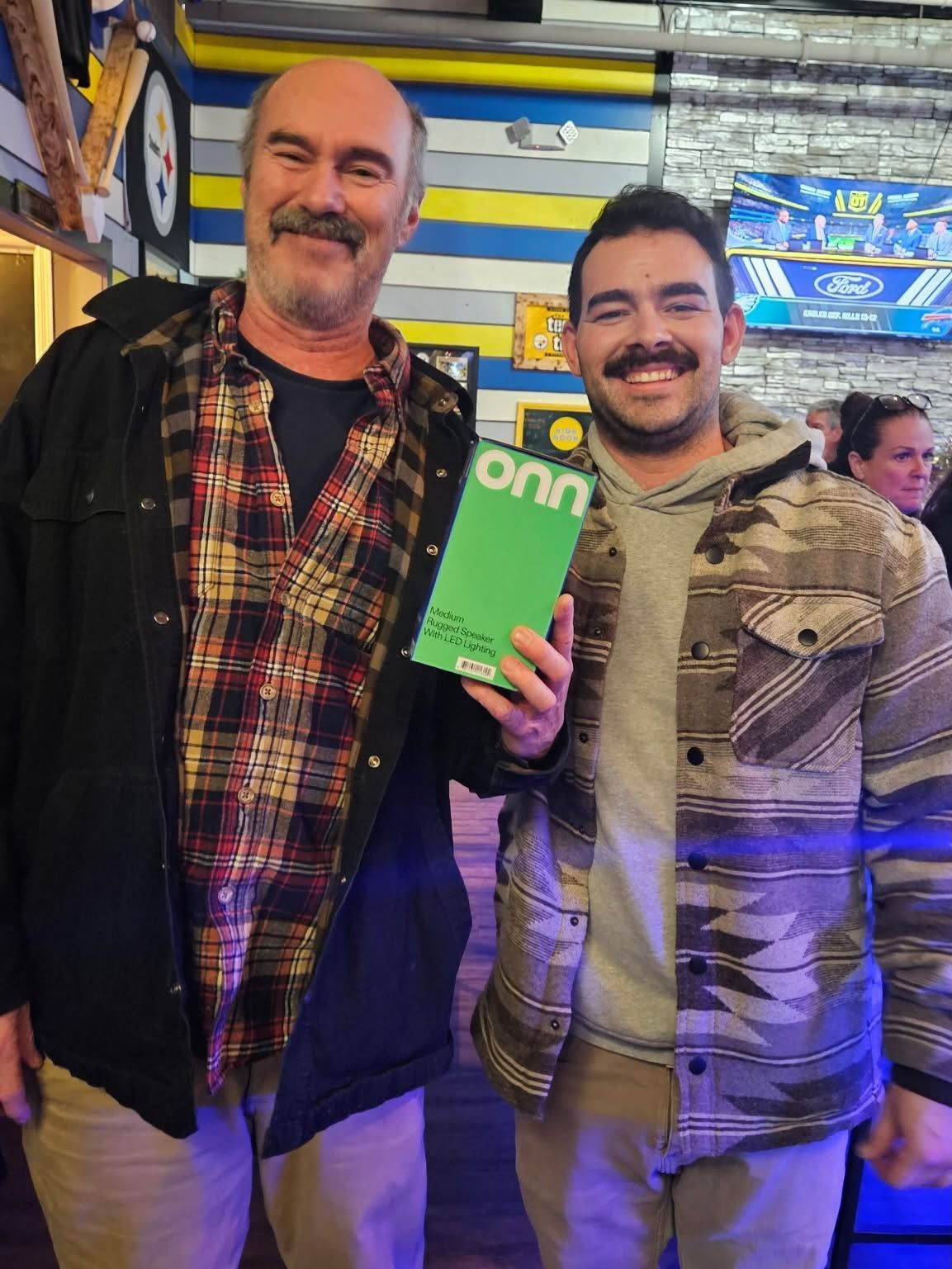 Two men smiling indoors, holding a green sign, with colorful lights and TV screens in the background