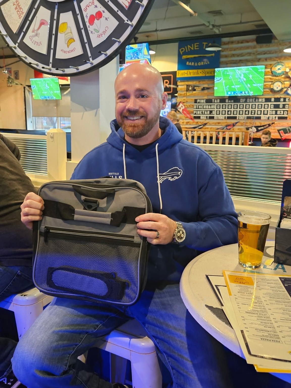Smiling person in a blue hoodie holding a black backpack at a restaurant table