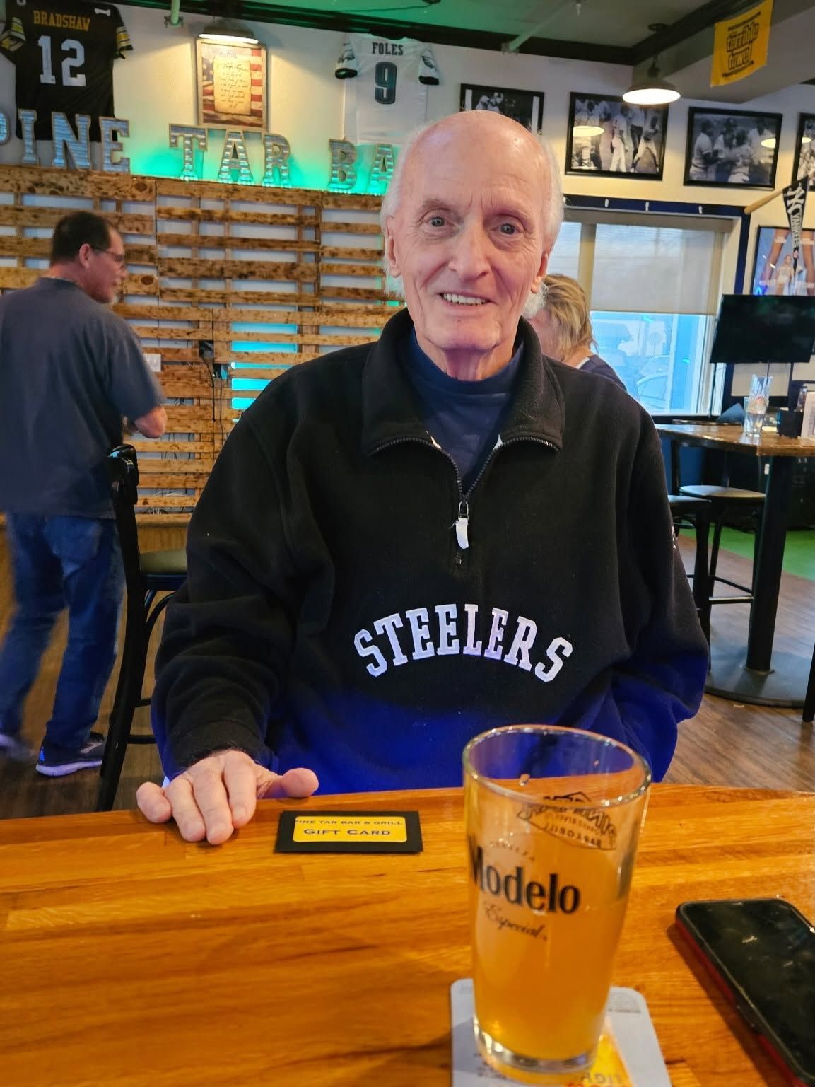 Man in a Steelers sweatshirt smiling at a bar with a beer in front of him