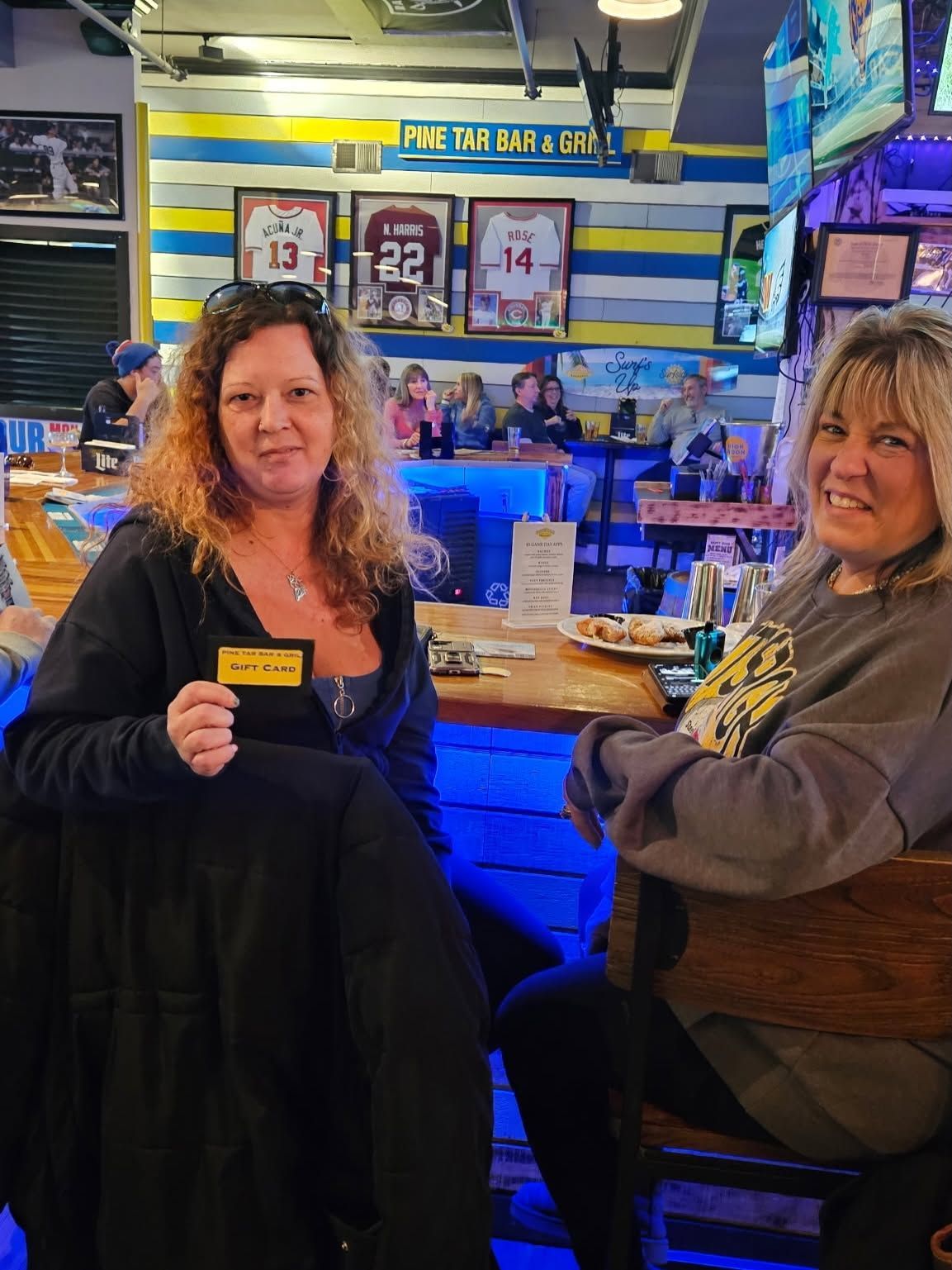 Two people seated at a bar, smiling in a neon-lit sports bar with TV screens and framed jerseys behind them.