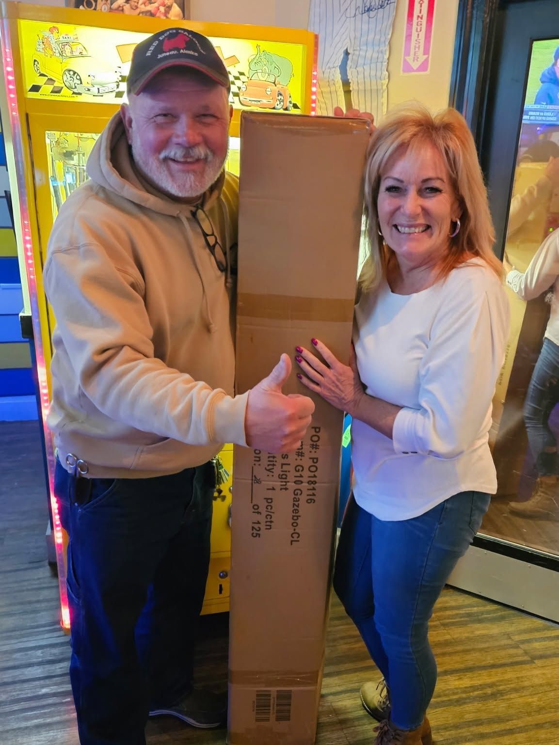 Two smiling people holding a tall cardboard box in a brightly lit store entrance