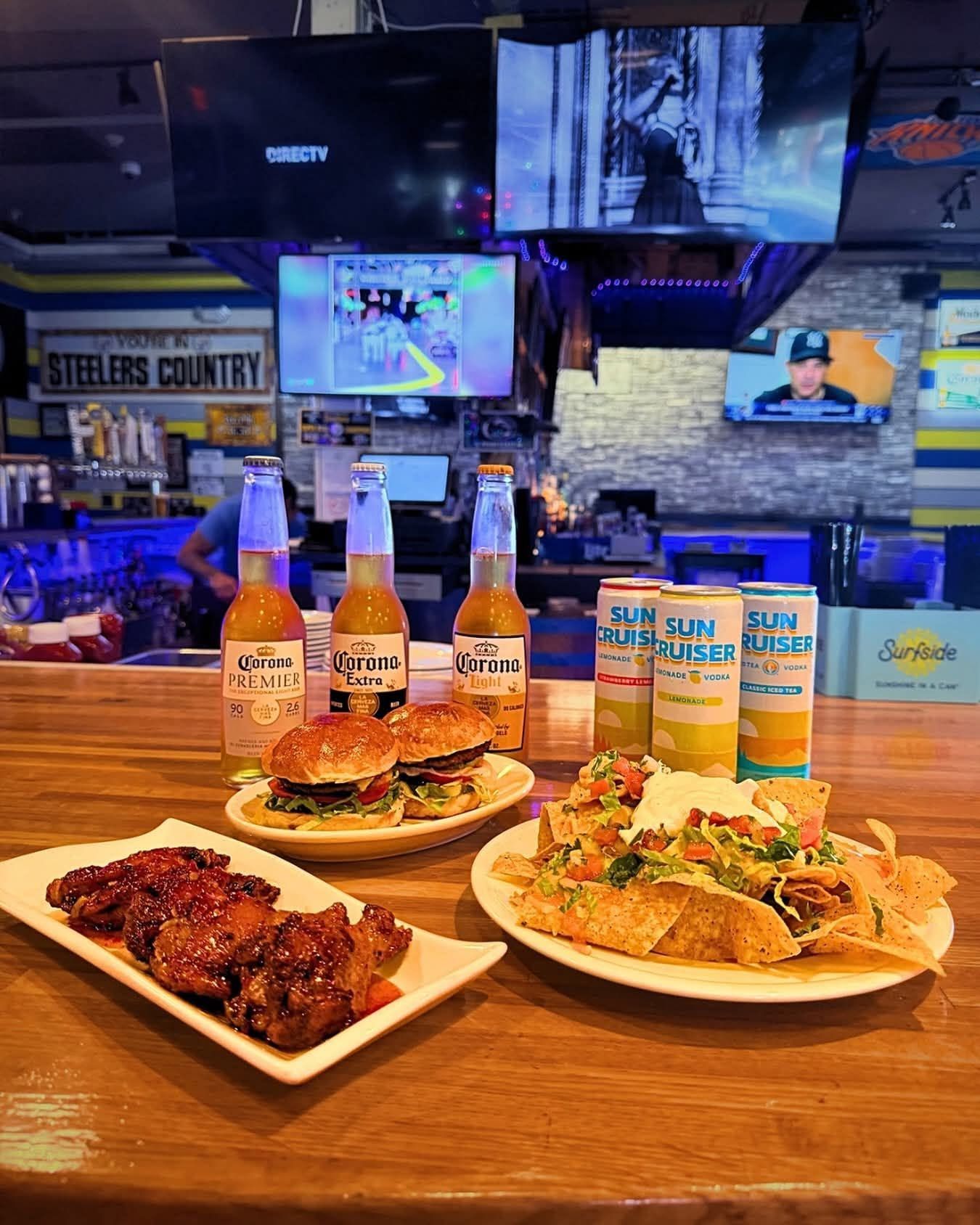 Bar food spread on a wooden table with wings, tacos, sliders, and bottles in a sports bar with TVs.