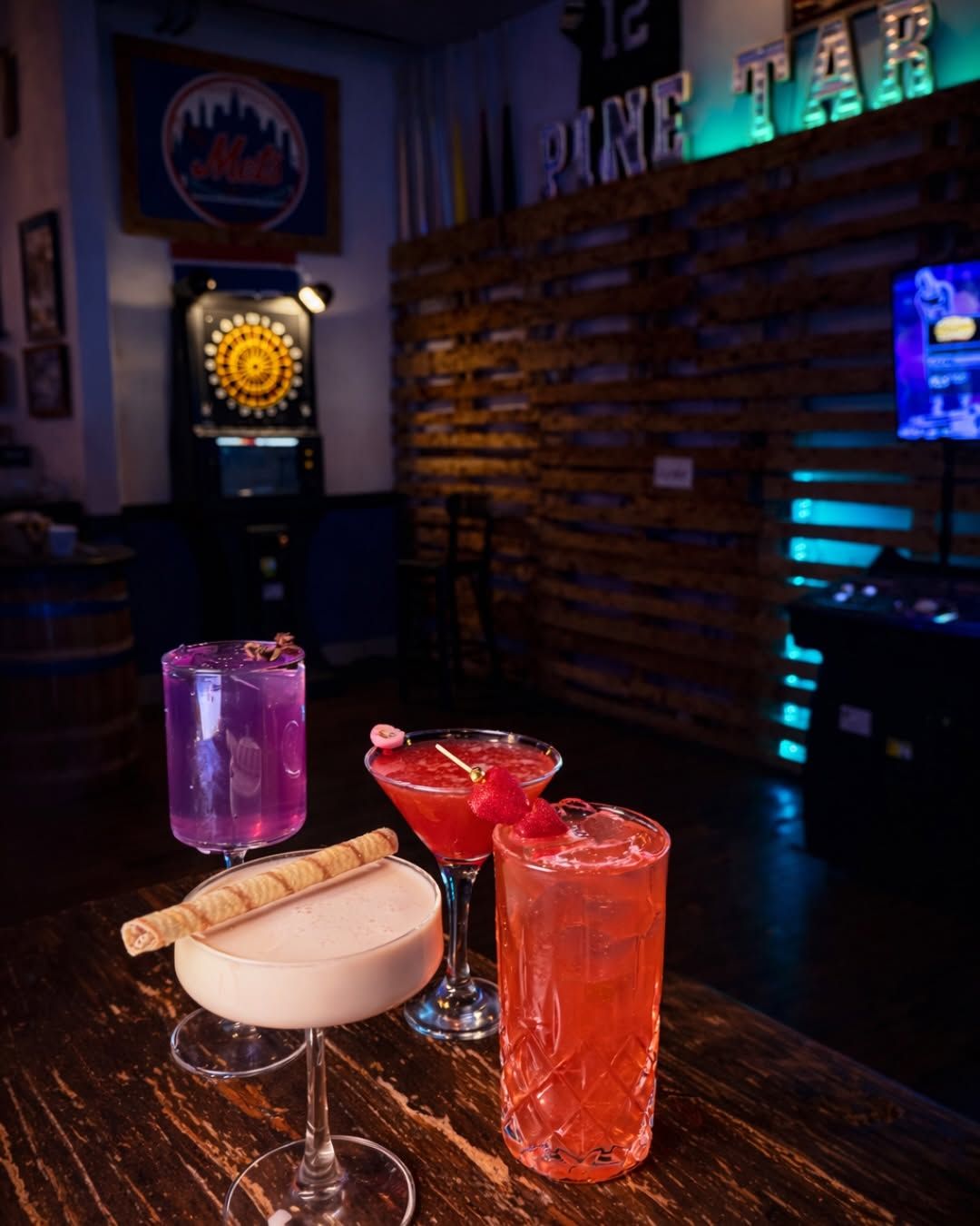Three colorful cocktails on a bar counter in a dimly lit lounge, with a neon bar sign in the background