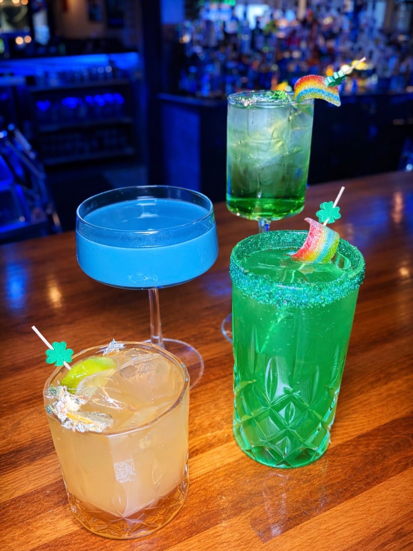 Four colorful tropical cocktails on a bar table, including blue, green, and orange drinks with fruit garnishes.