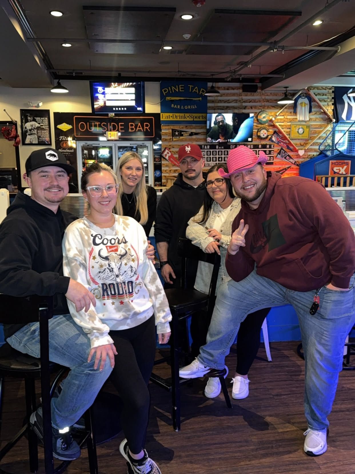 Group of people smiling and posing in a lively bar with neon signs and TVs in the background