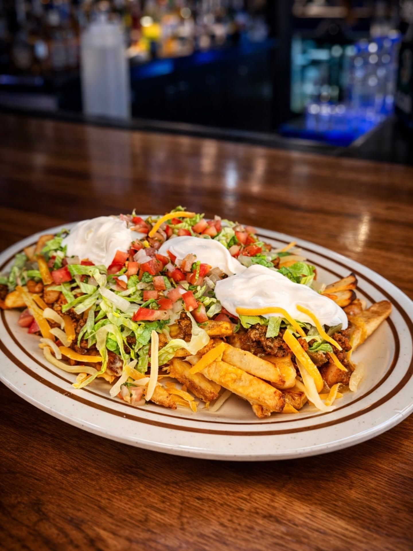 Loaded fries topped with shredded lettuce, tomatoes, sour cream, and melted cheese on a plate at a bar counter