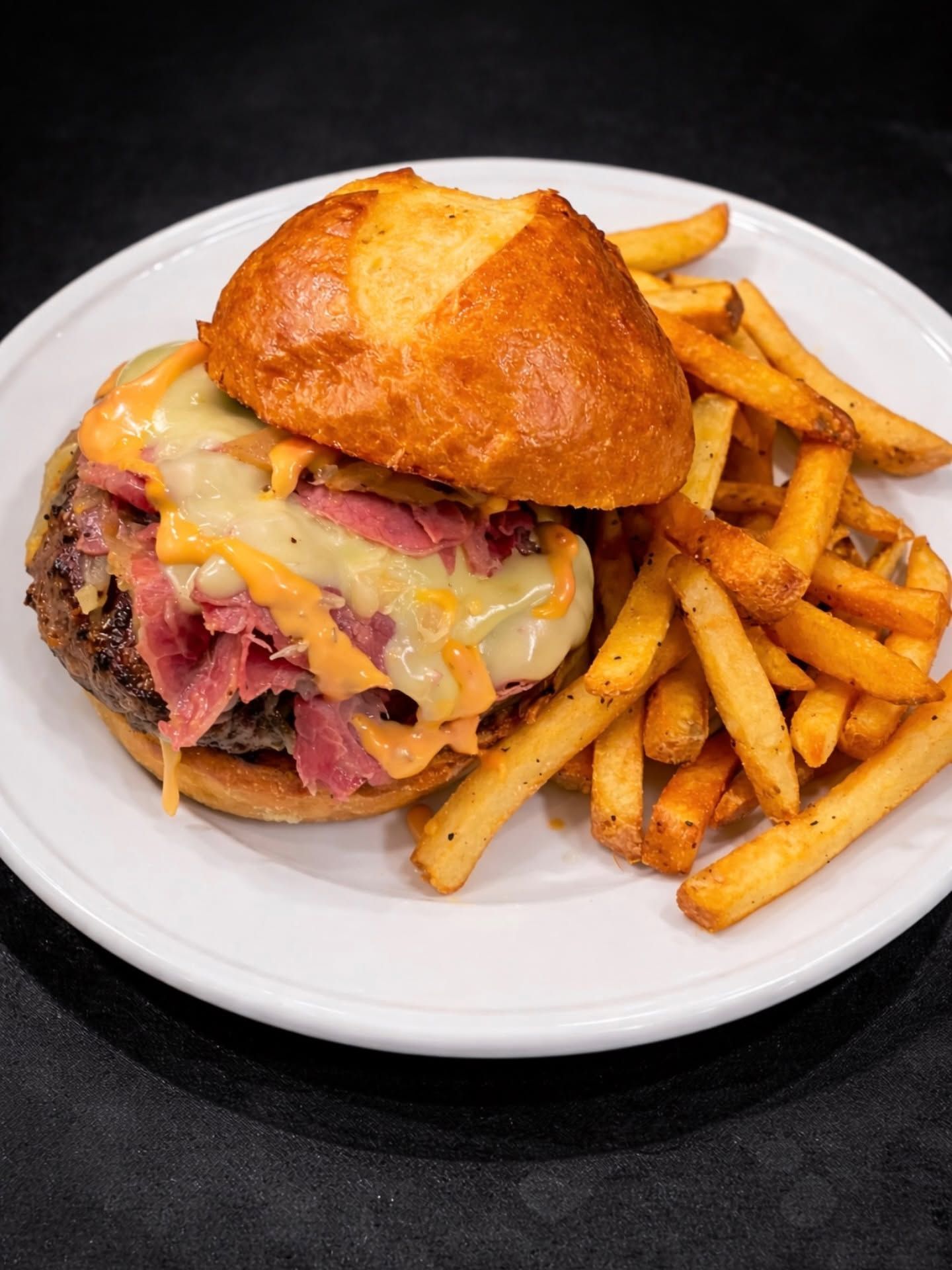 Burger with melted cheese and fries on a white plate on a dark surface