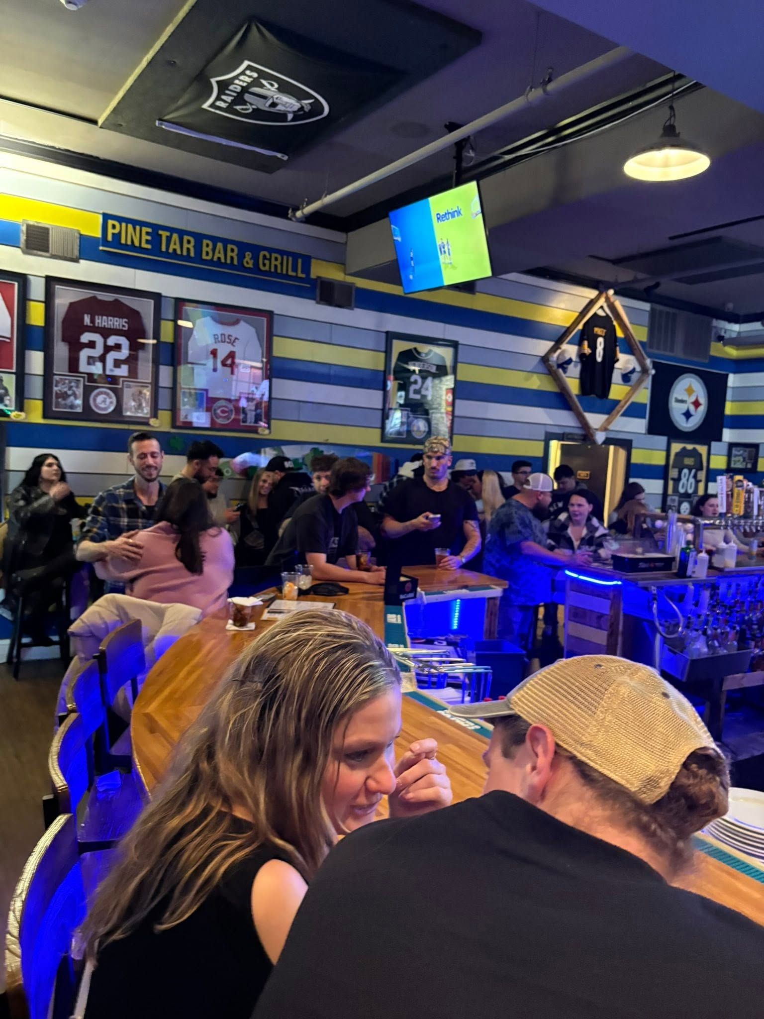 Crowded sports bar with patrons seated at a blue-lit counter, TVs above, and football memorabilia on the walls