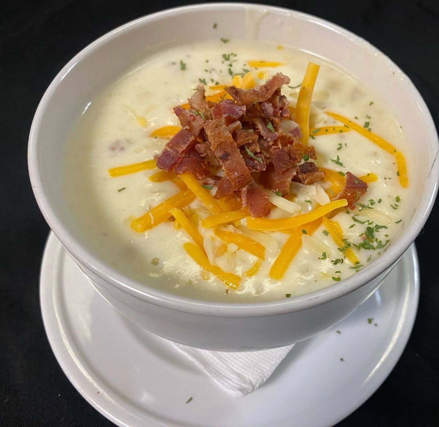 Creamy soup topped with shredded cheddar, bacon bits, and herbs in a white bowl and saucer