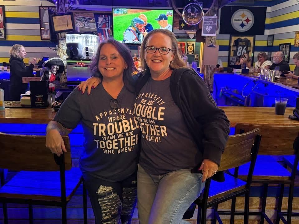 Two women in matching shirts pose smiling at a dimly lit bar with blue lights and TVs in the background