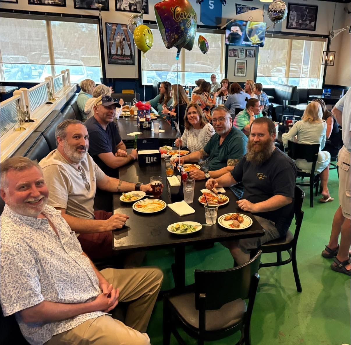 Group of people seated around tables in a casual restaurant, eating meals and smiling.