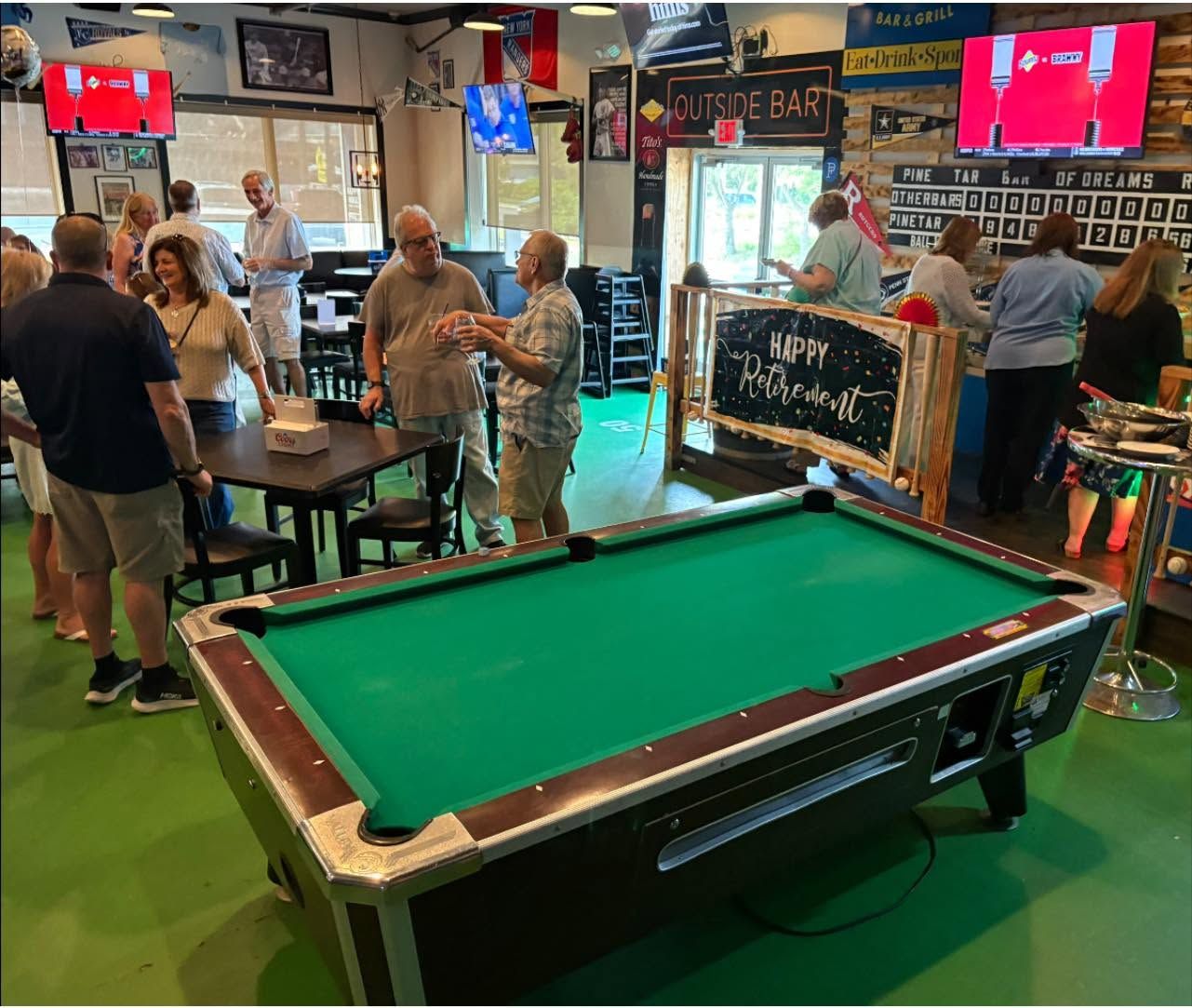Green billiards table in a busy bar with patrons standing around and TVs showing sports