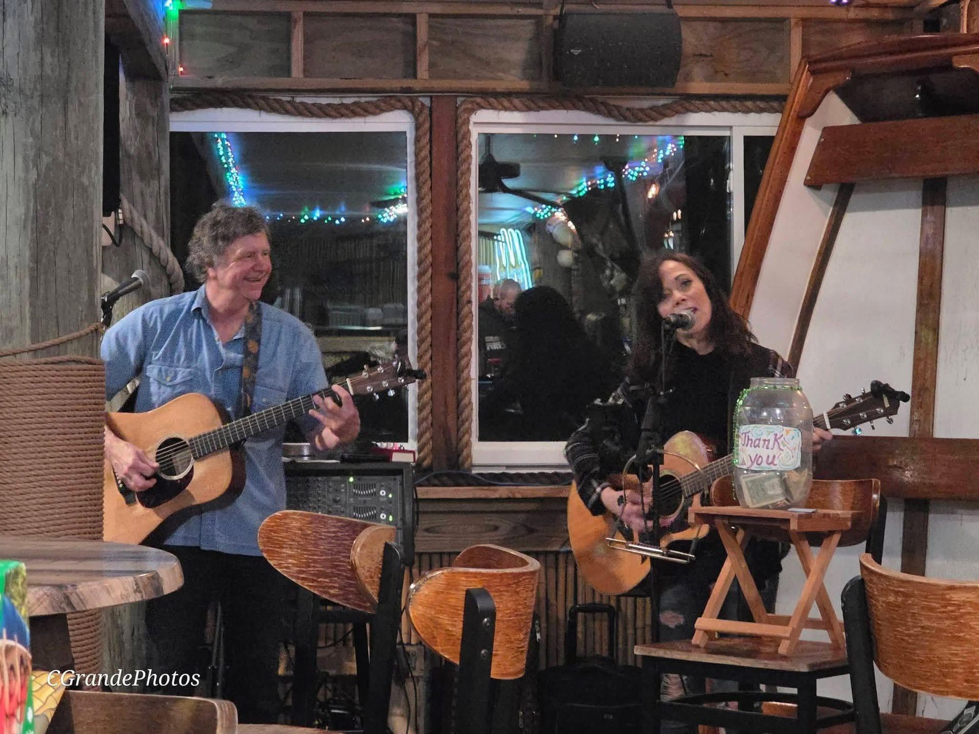 Two musicians playing acoustic guitars in a cozy café by large windows at night