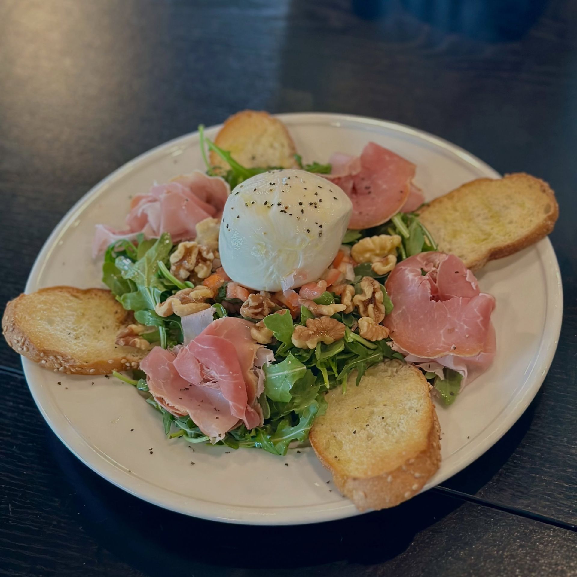 Salad with burrata, prosciutto, walnuts, and toasted bread on a white plate