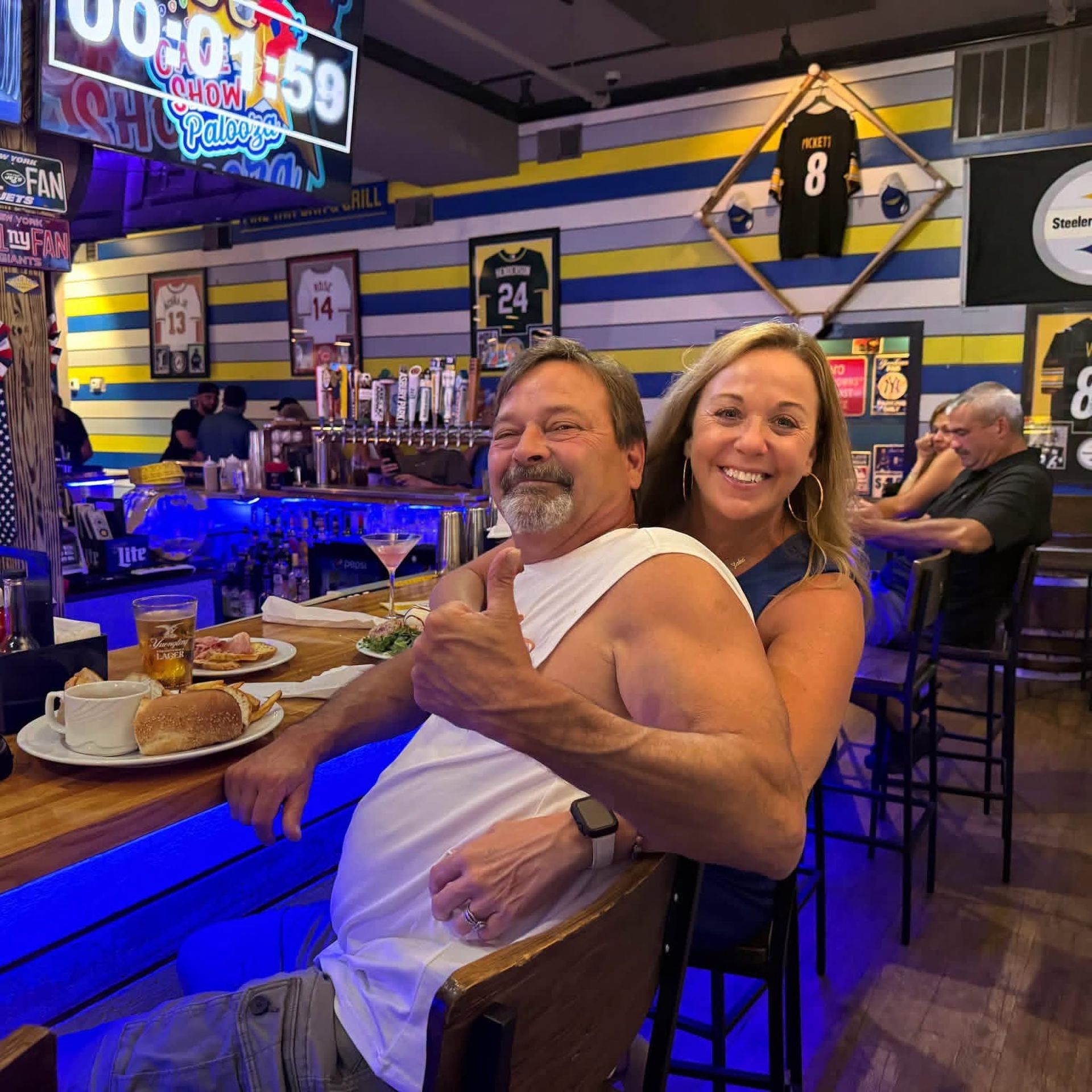 Couple smiling at a bar counter with drinks, in a lively sports bar with neon lights and jerseys.