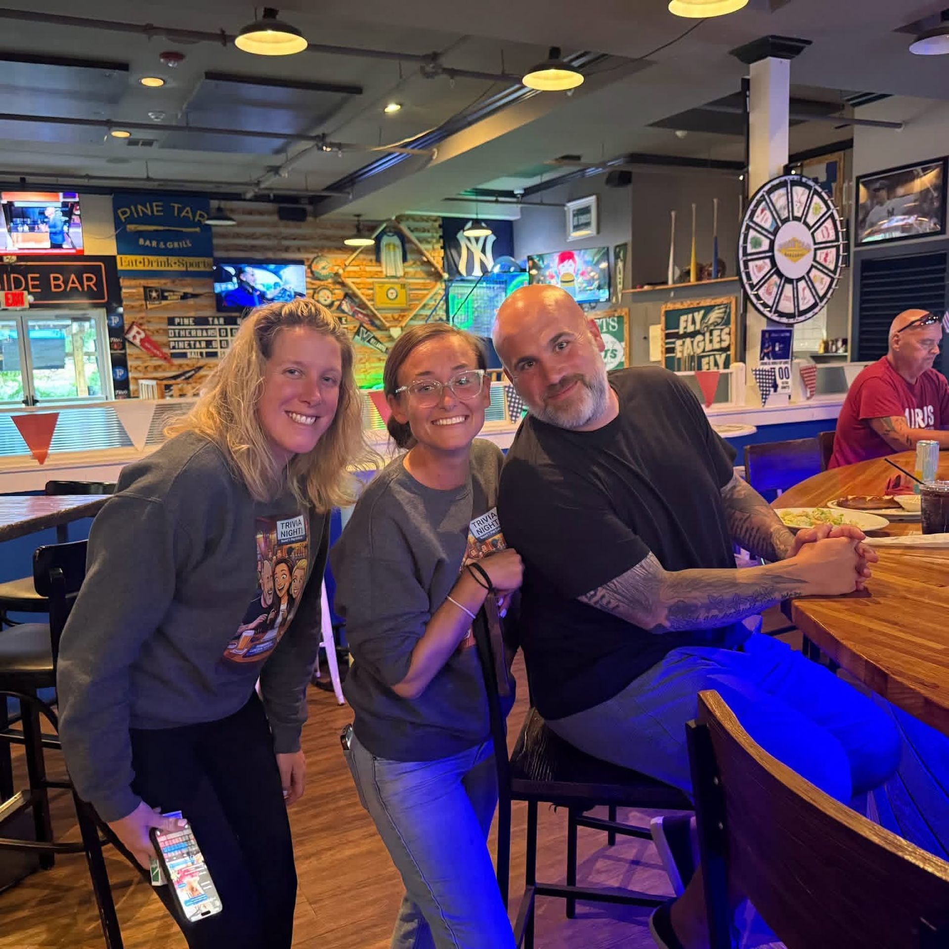 Three people smiling at a bar, leaning on stools under colorful neon lights.