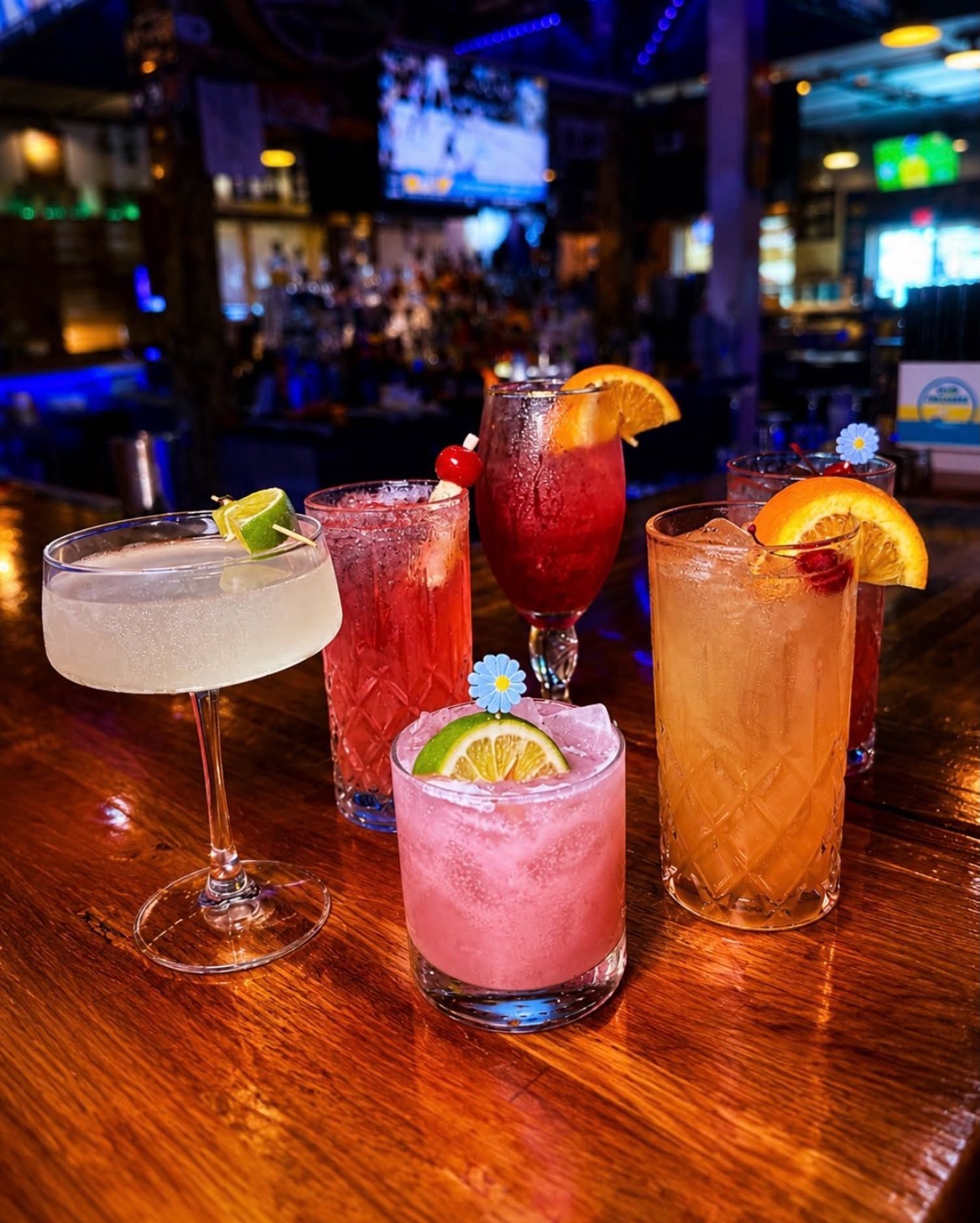 Four colorful cocktails on a bar counter with a blurred nightlife background.
