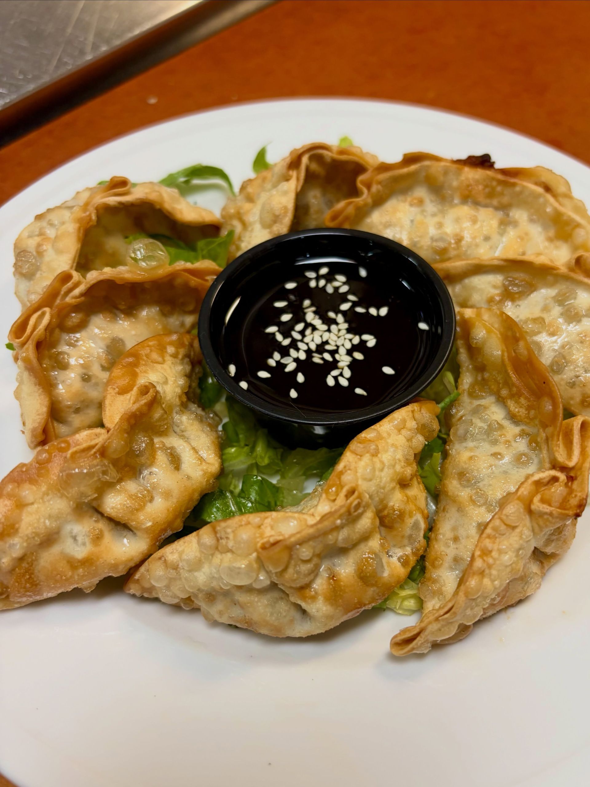 Plate of golden fried dumplings arranged around a black dipping sauce bowl, garnished with sesame seeds