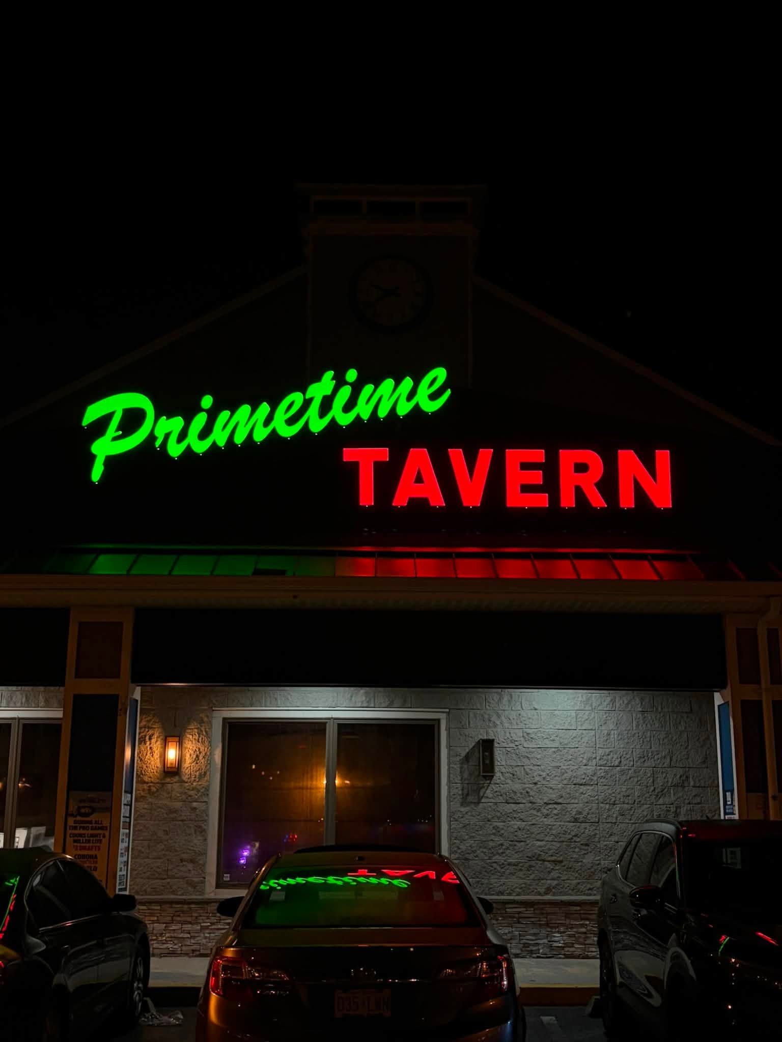 Neon sign for Primetime Tavern above a dimly lit storefront at night, with parked cars in front.